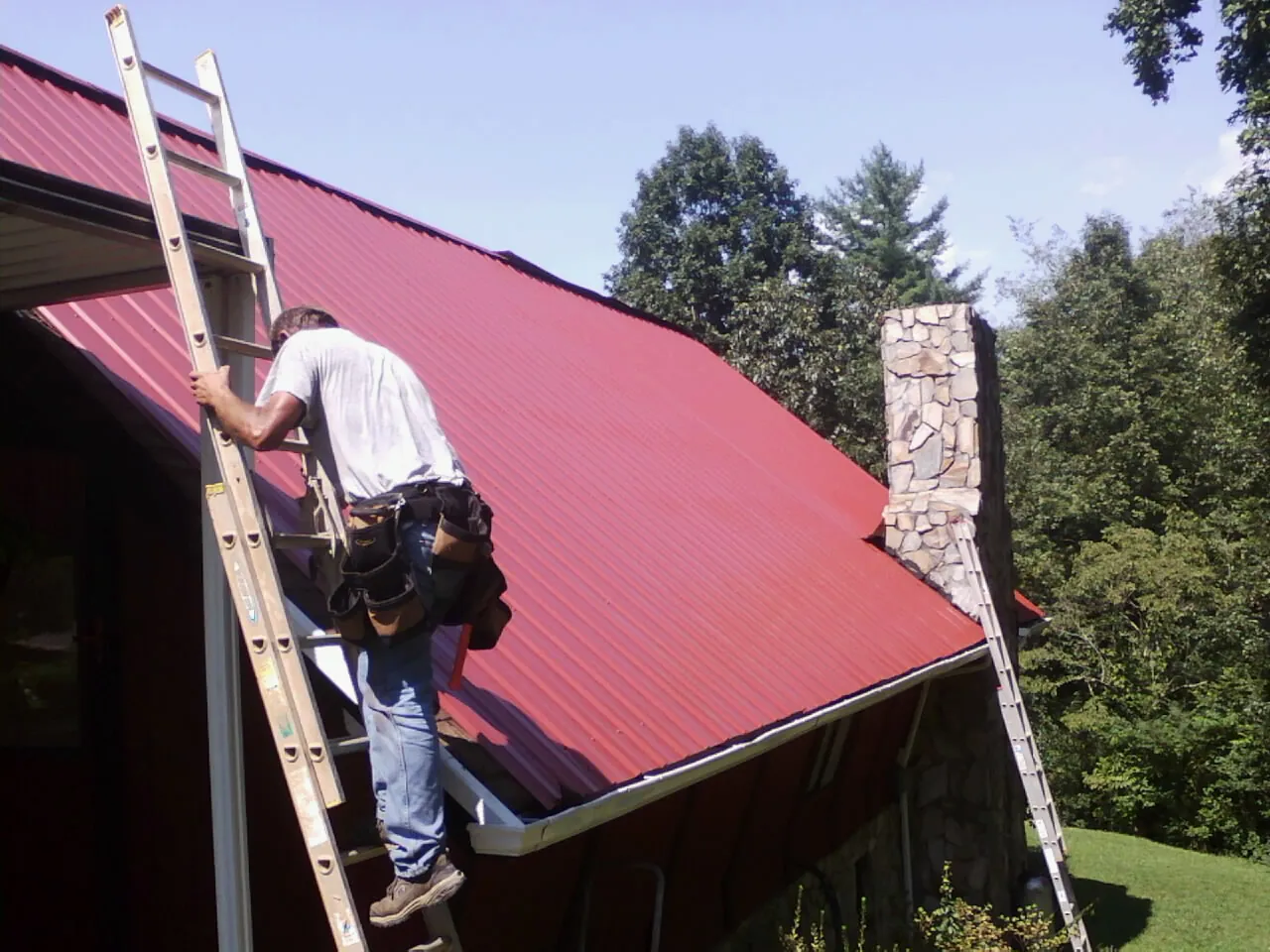 Roofer on a ladder beside a red metal roof and stone chimney — Soffit & Fascia Repair by Roofing Brothers in Caneyville